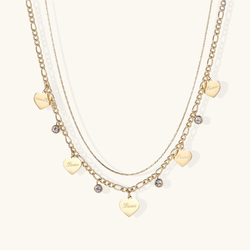 Gold layered necklace set with engraved heart pendants and crystal accents on cream background